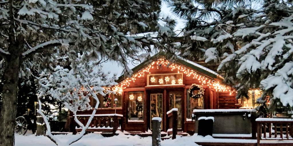 A single story wood cabin with small lights along the trim, two big windows and double glass doors on the front. A wood deck extends in front. The whole scene is covered in snow and trees with snow are in the foreground