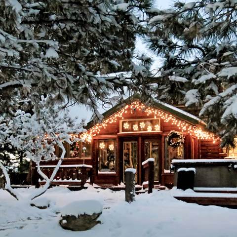 A single story wood cabin with small lights along the trim, two big windows and double glass doors on the front. A wood deck extends in front. The whole scene is covered in snow and trees with snow are in the foreground